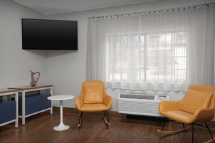 Quarto com televisão de tela plana na parede, duas cadeiras amarelas, uma pequena mesa branca, um banco azul e uma janela com cortinas leves.