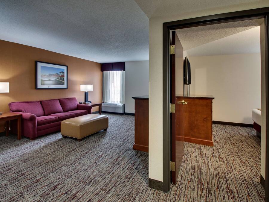 Quarto de hotel com sofá, puf, duas mesas de cabeceira com luminárias, escrivaninha de madeira e uma grande janela com cortinas.