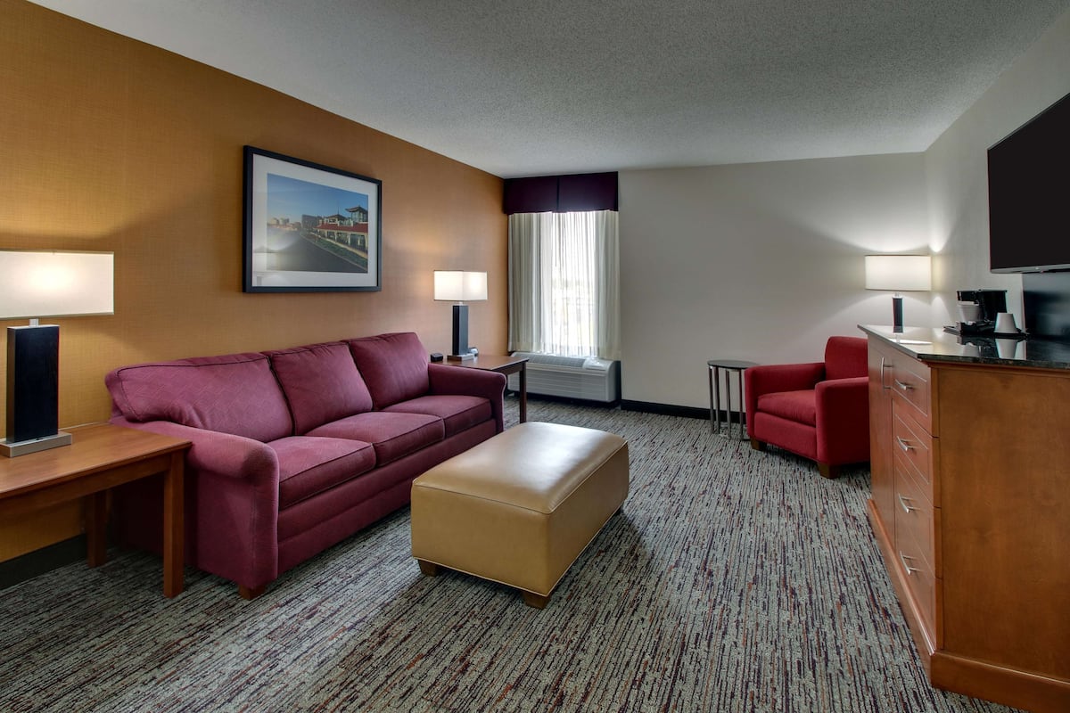 A hotel room with a red sofa, a beige ottoman, a wooden desk with a lamp, a small table, a red armchair, a TV, and a window with curtains.