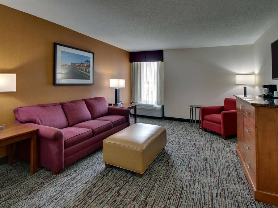 A hotel room with a red sofa, a beige ottoman, a wooden desk with a lamp, a small table, a red armchair, a TV, and a window with curtains.