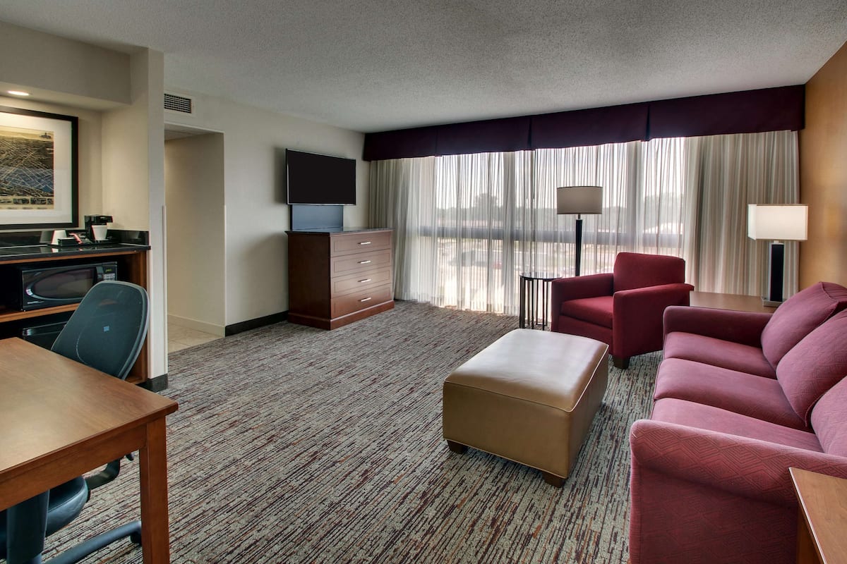 A hotel room with a wooden desk, a red sofa, a brown ottoman, a TV on a stand, a dresser, and a large window with sheer curtains.