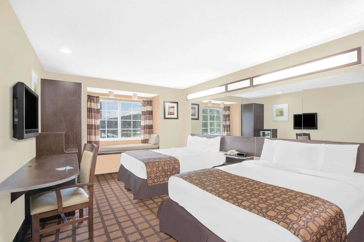 A hotel room with two beds, a desk, a chair, a TV, a window with curtains, and a view of the outdoors.