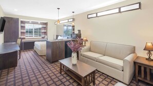 Ein Hotelzimmer mit einer Sitzecke, einem kleinen Tisch mit einer Vase, einem Schreibtisch, einem Stuhl, einer Küchenzeile und einem Fenster mit Vorhängen.