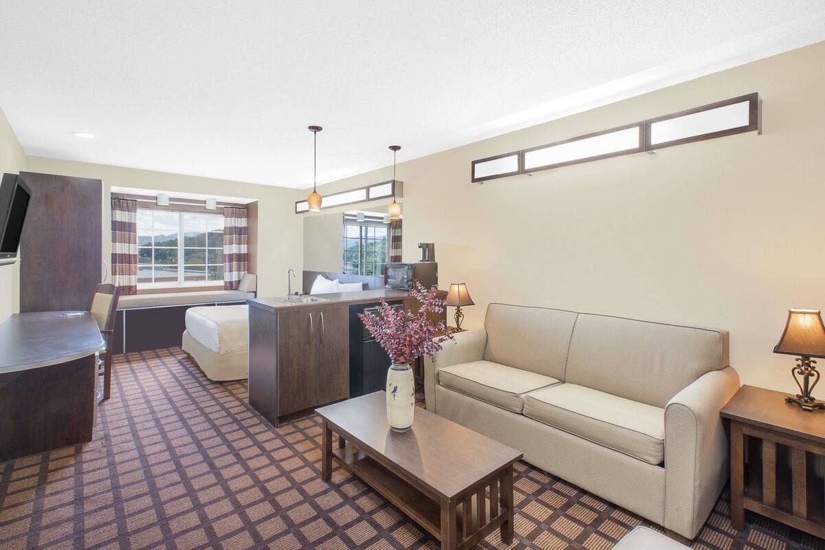 A hotel room with a sofa, a small table with a vase, a desk, a chair, a kitchen area, and a window with curtains.
