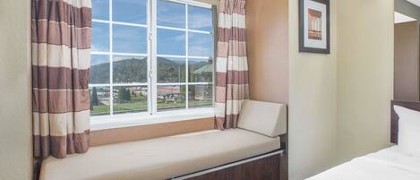 A hotel room with a bed, a window with curtains, a bench, and a framed picture on the wall.