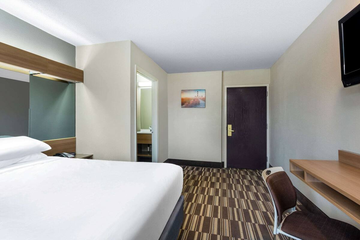 A hotel room with a bed, a desk, a chair, a TV, a door, and a painting on the wall.