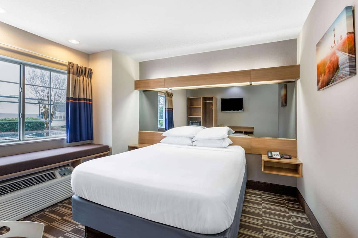 A hotel room with a large bed, a bedside table, a mirror, a TV, and a window with curtains.