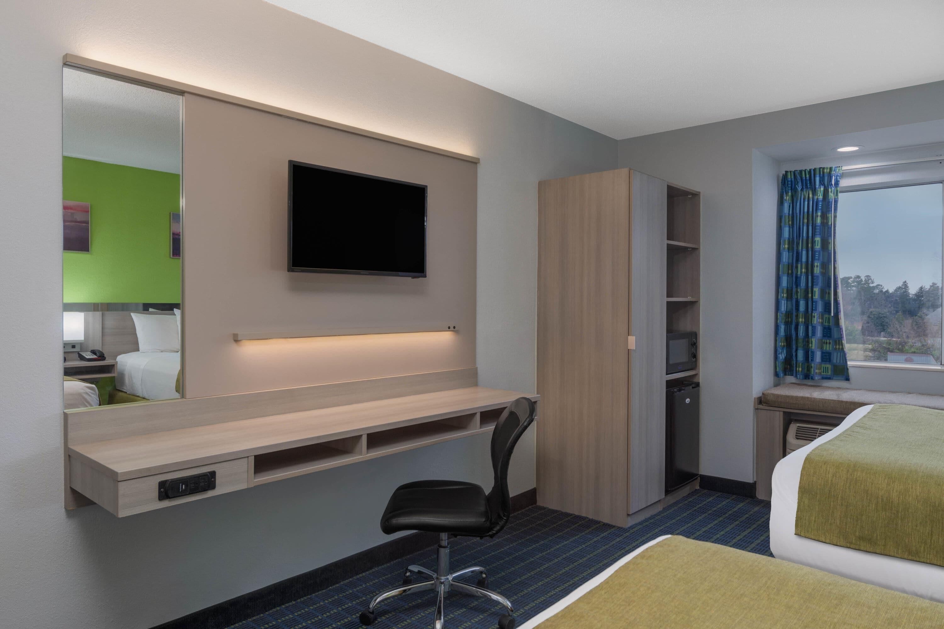 Une chambre d’hôtel équipée d’un bureau, d’une chaise, d’un lit, d’une télévision, d’une armoire et d’une fenêtre avec des rideaux.