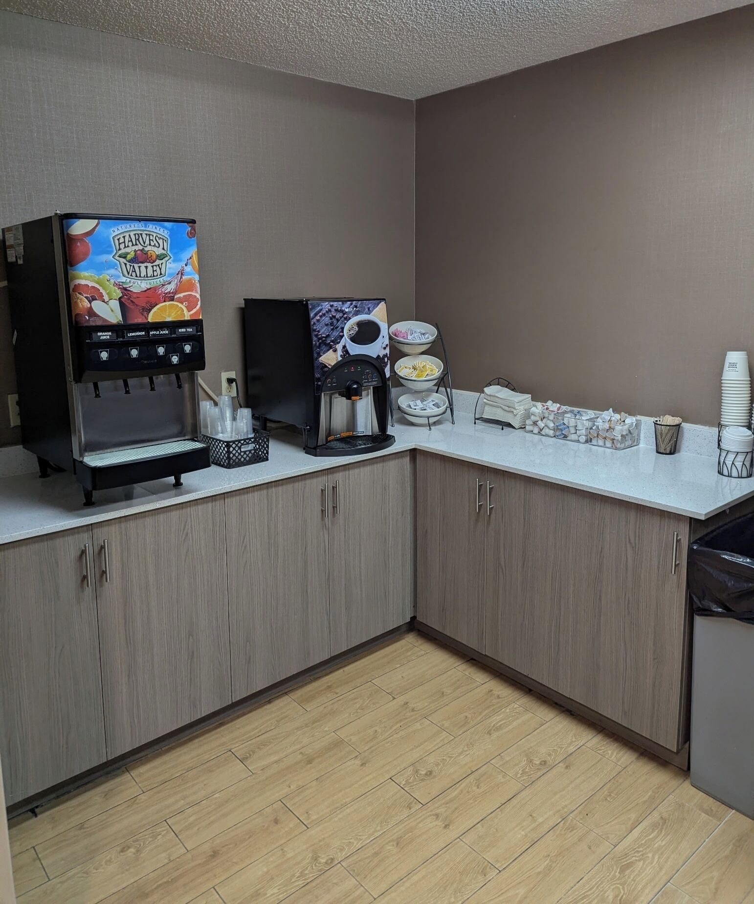 A kitchenette with a coffee machine, a fruit juice dispenser, and a stack of white cups.