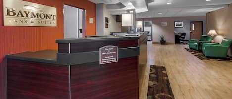 Un mostrador de recepción con un cartel que indica el nombre del hotel y sus servicios, un área de espera con sillas verdes y suelo cubierto de alfombra.