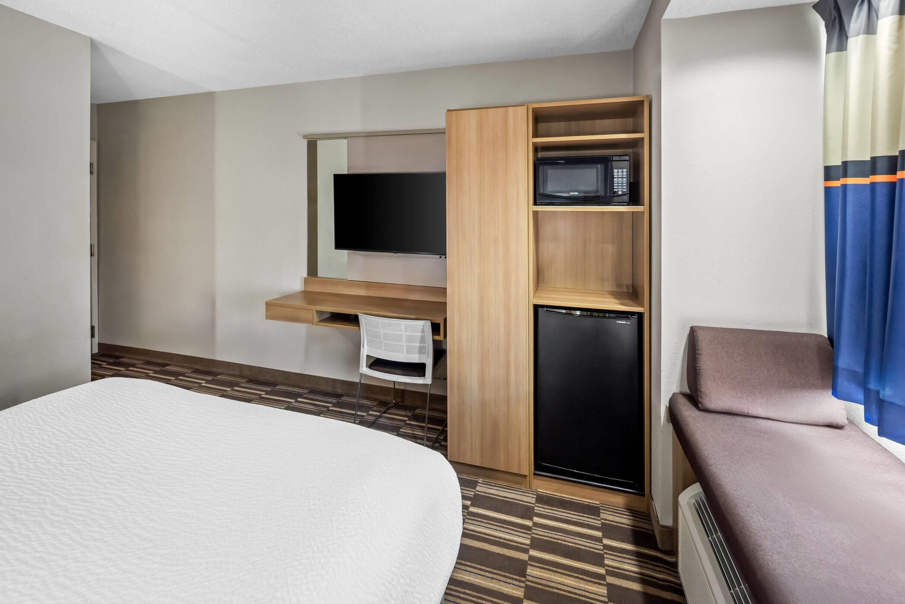 A hotel room with a bed, a desk with a flat-screen TV, a refrigerator, a microwave, and a seating area with a cushion.