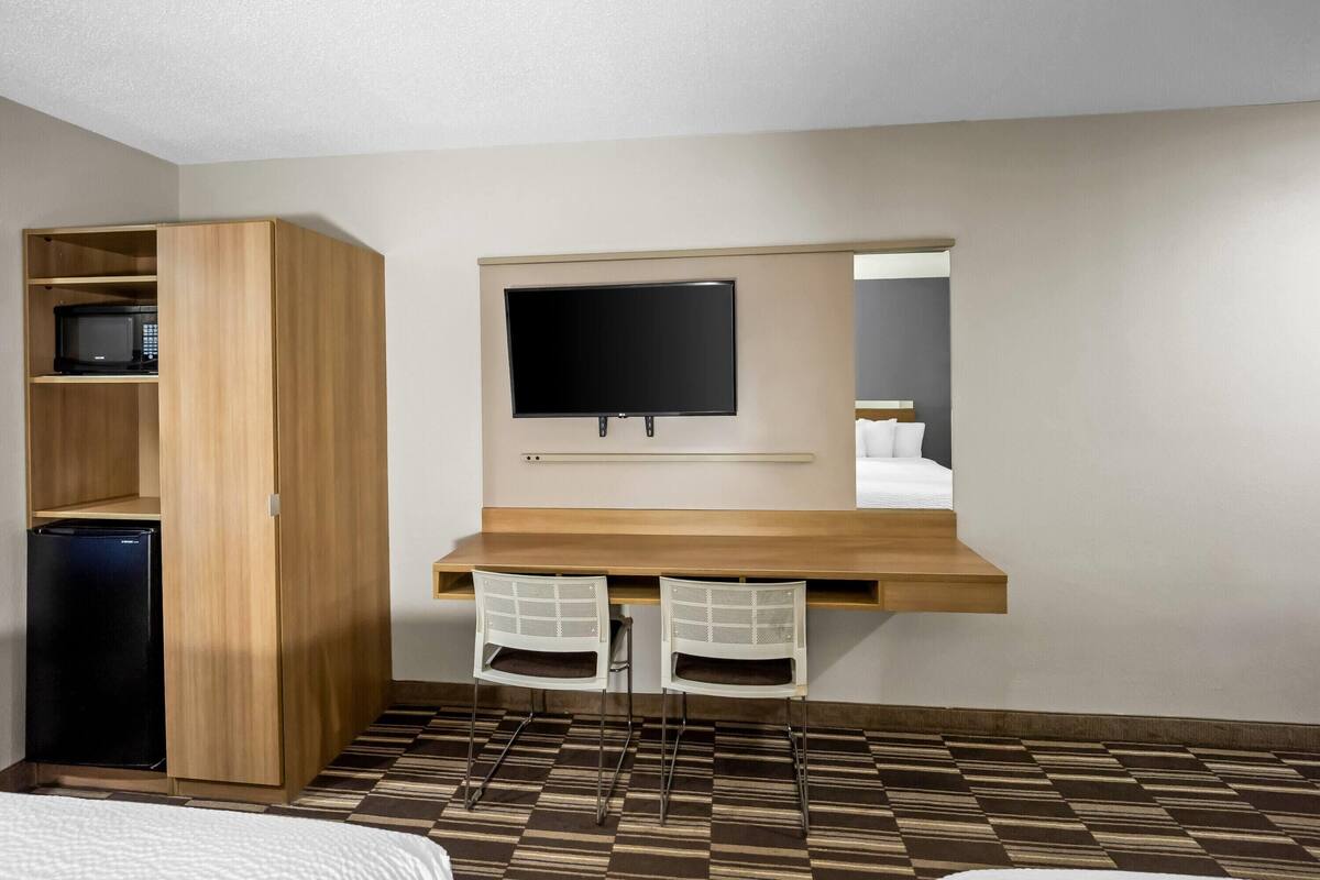 A hotel room with a wooden desk, two chairs, a television, a minibar, and a patterned floor.