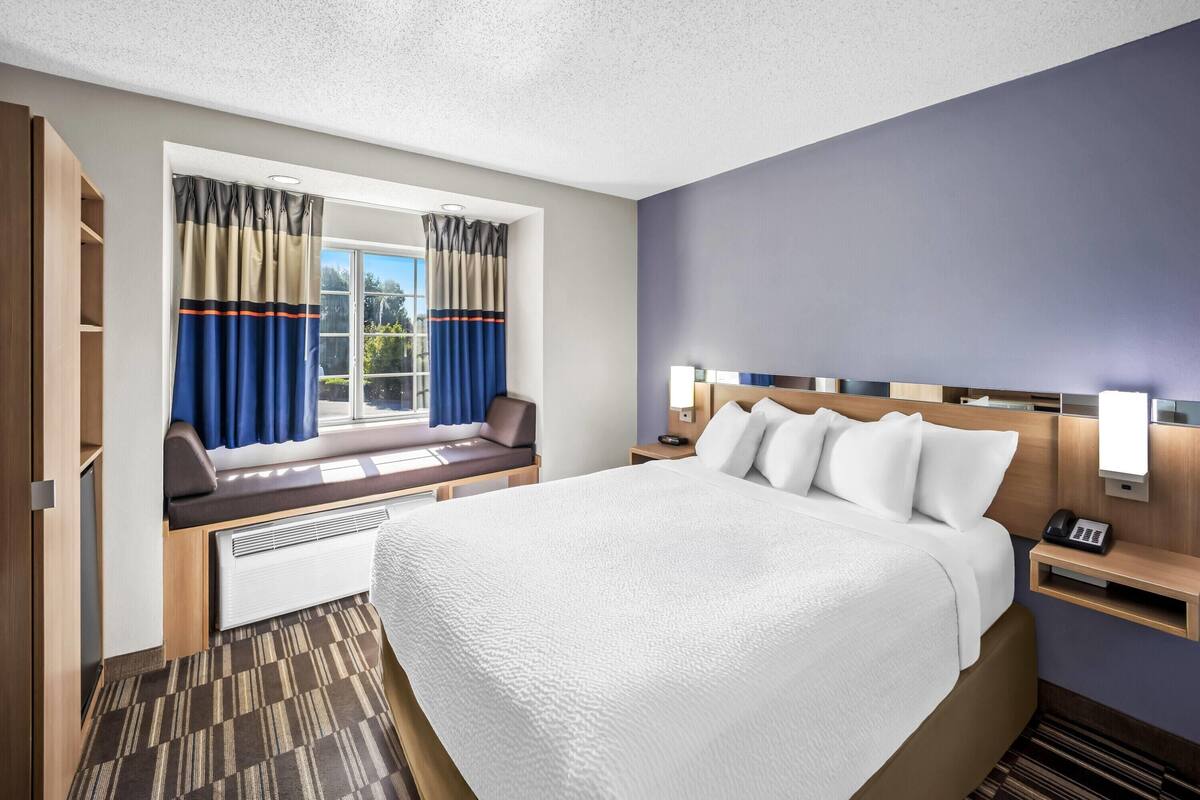 A hotel room with a large bed, a window seat with curtains, a bedside table with a lamp, and a wall-mounted phone.