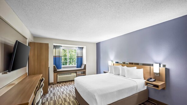 A hotel room with a large bed, a desk with a TV, a window with curtains, and a nightstand with a phone and lamp. - Microtel Inn by Wyndham Greensboro (Greensboro)