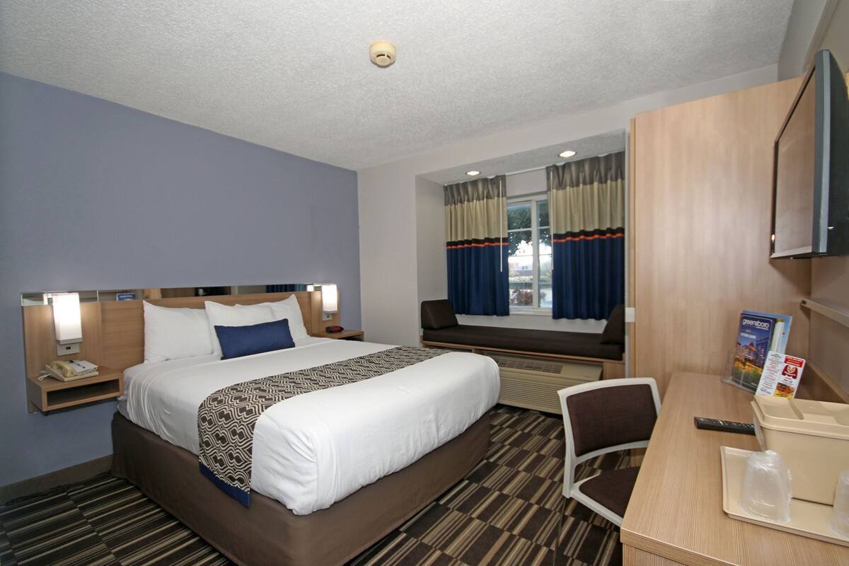 A hotel room with a large bed, bedside tables, a desk, a chair, a TV, and a window with curtains.