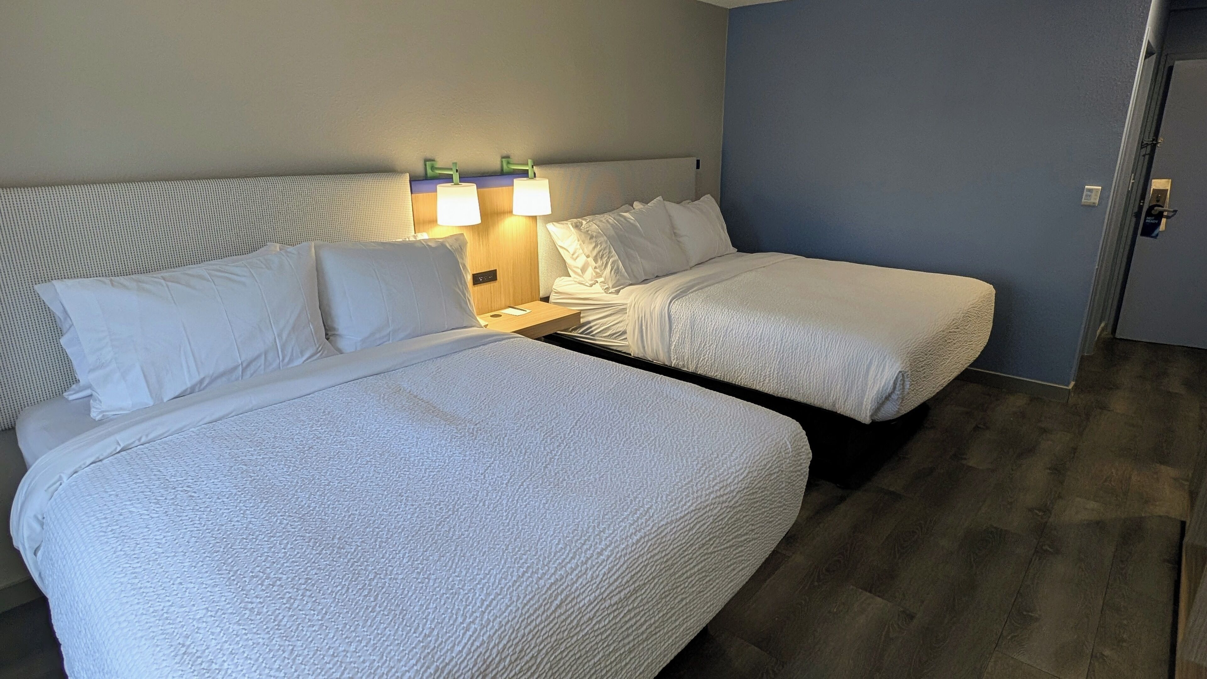 Habitación de hotel con dos camas, una mesita de noche con lámparas, un cabecero de madera y una puerta a la derecha.