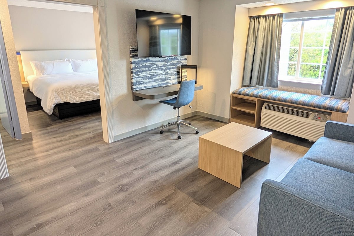 Habitación de hotel con cama, escritorio y silla, sofá, televisión y ventana con cortinas.