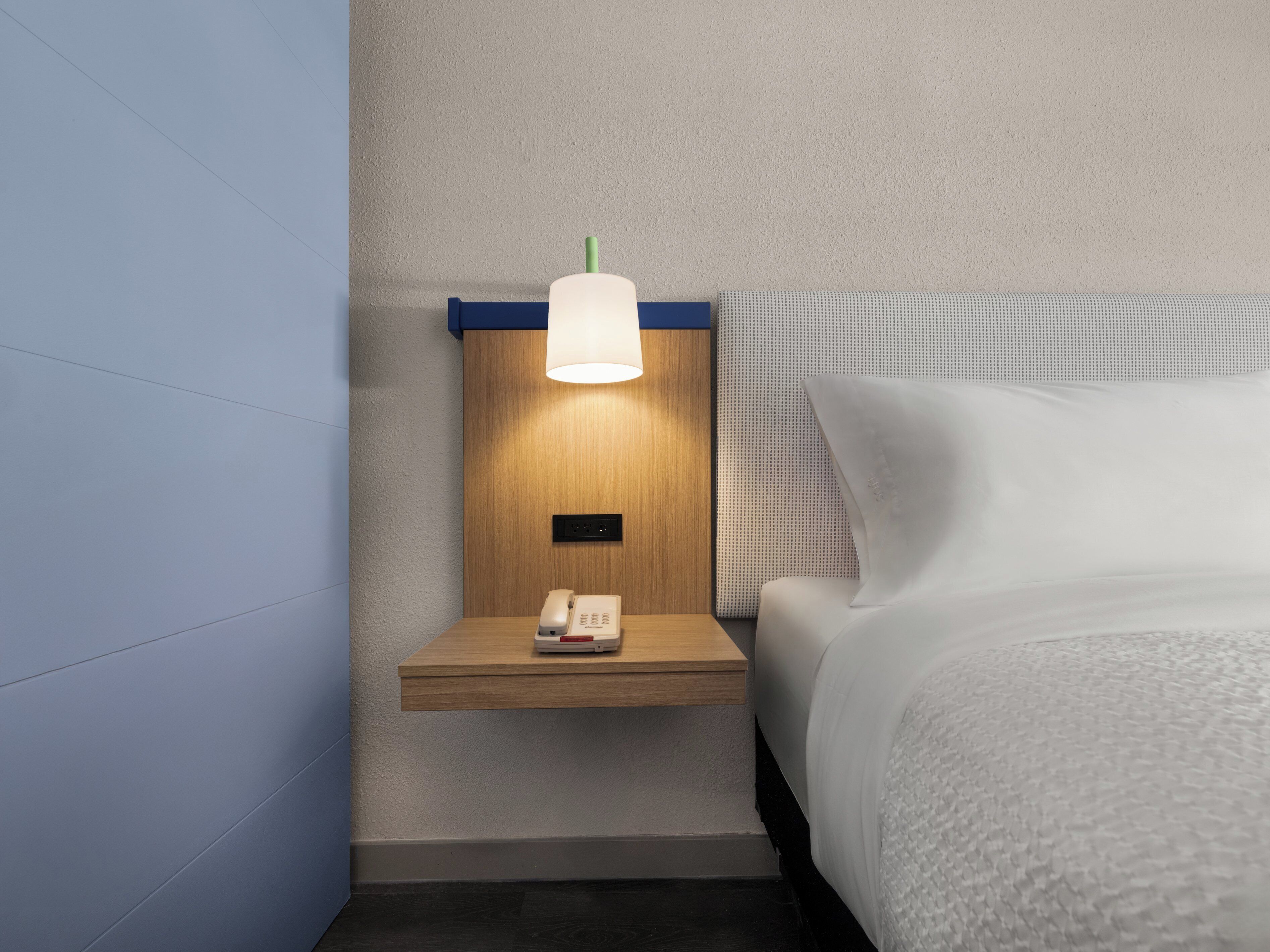Habitación de hotel con mesita de noche de madera, lámpara, teléfono y cama con ropa blanca.
