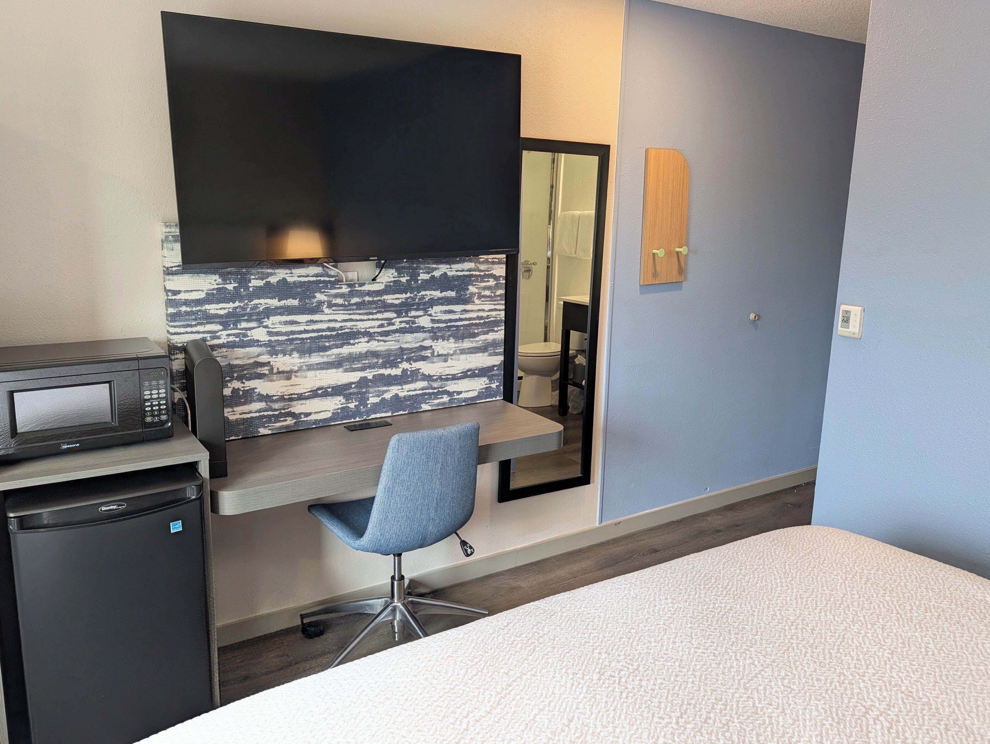 A hotel room with a desk, a microwave, a chair, a large mirror, and a small refrigerator.