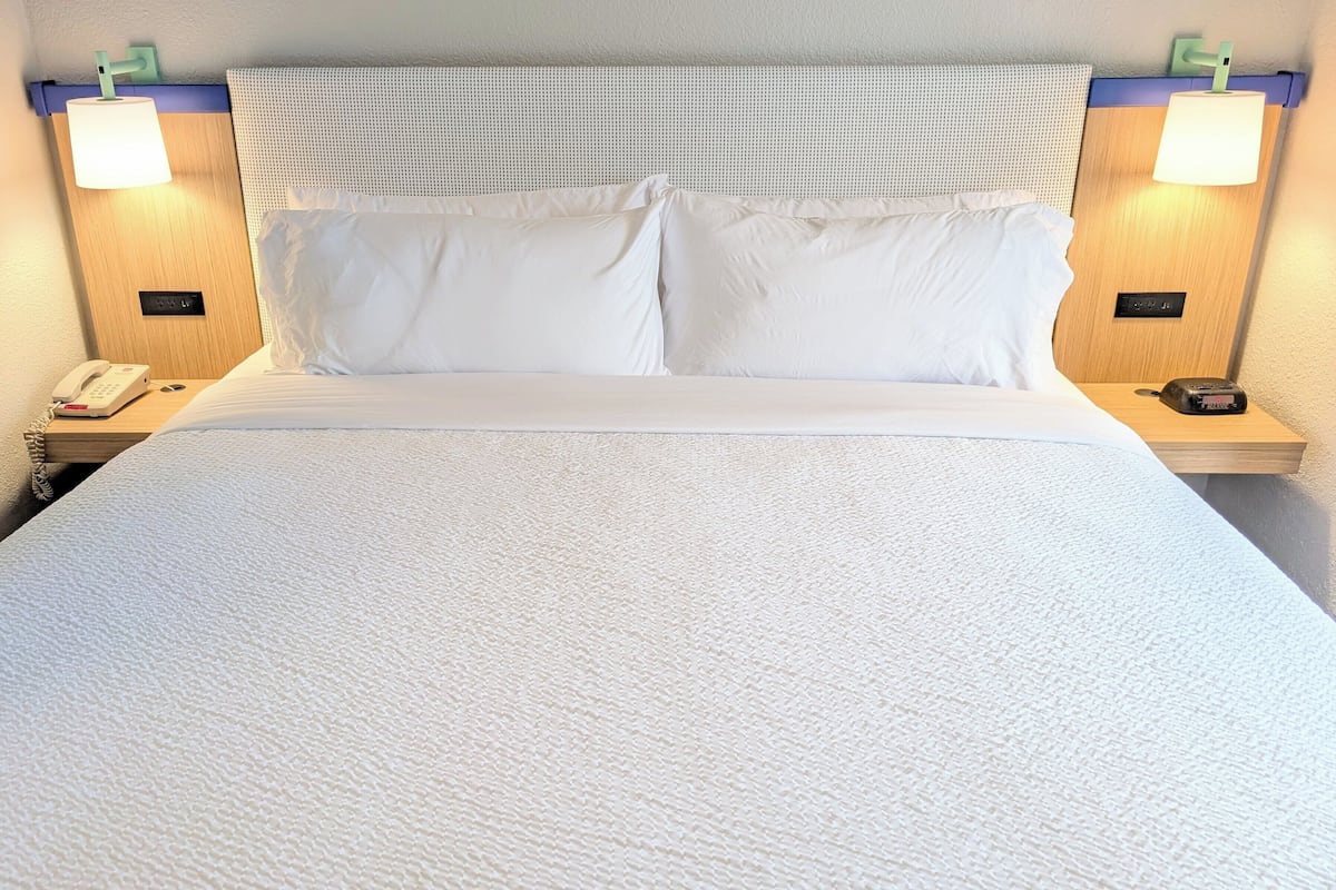 Una cama bien hecha con sábanas blancas, dos lámparas de noche, un teléfono y un reloj pequeño en la mesita de noche.