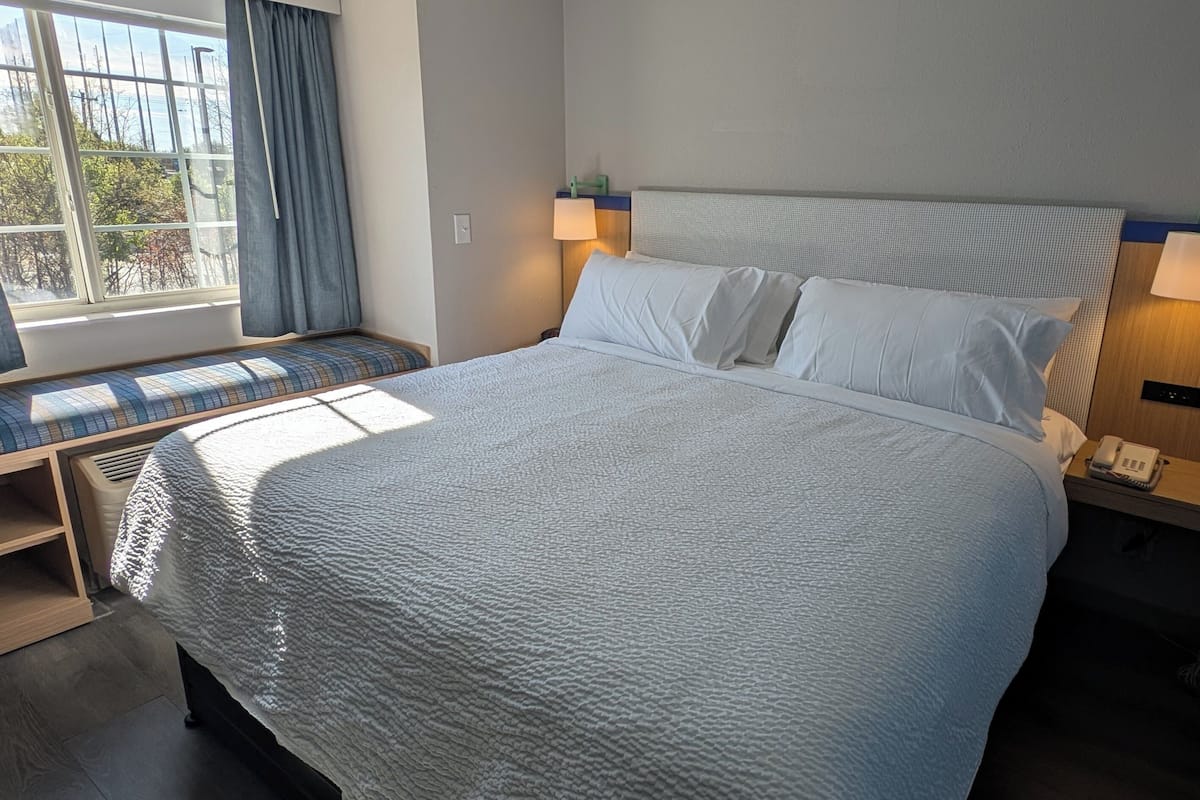 Una cama bien hecha con un edredón blanco texturizado, un banco con cojín escocés, una lámpara de noche y un teléfono en la mesita.