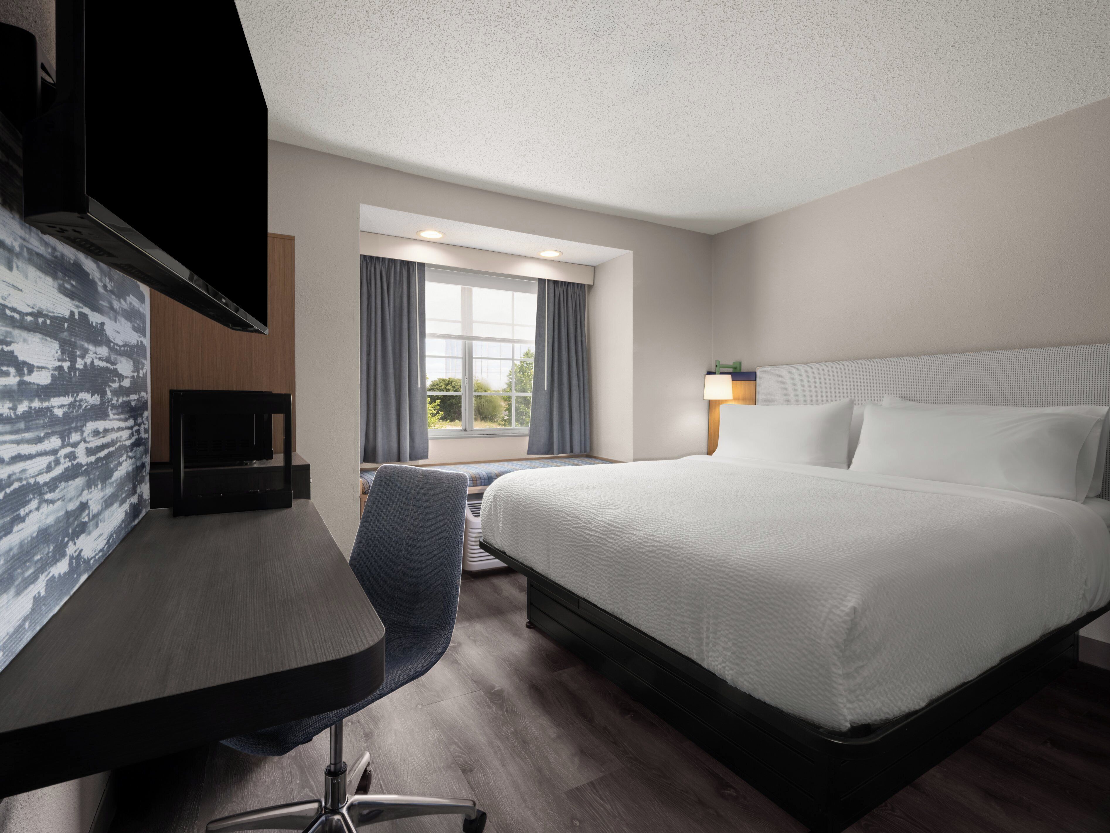 Une chambre d’hôtel avec un lit, un bureau et une chaise, une télévision et une fenêtre avec des rideaux.