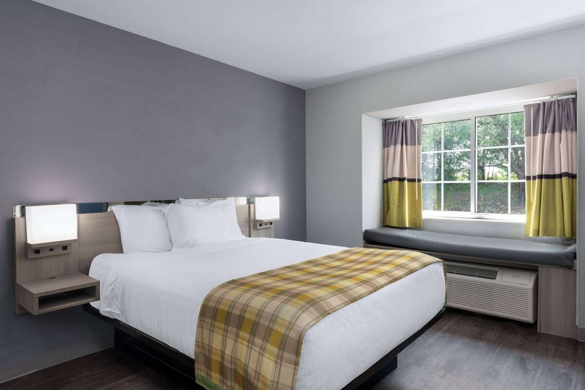 Una habitación de hotel con cama, lámparas de noche, ventana con cortinas y un banco empotrado debajo de la ventana.