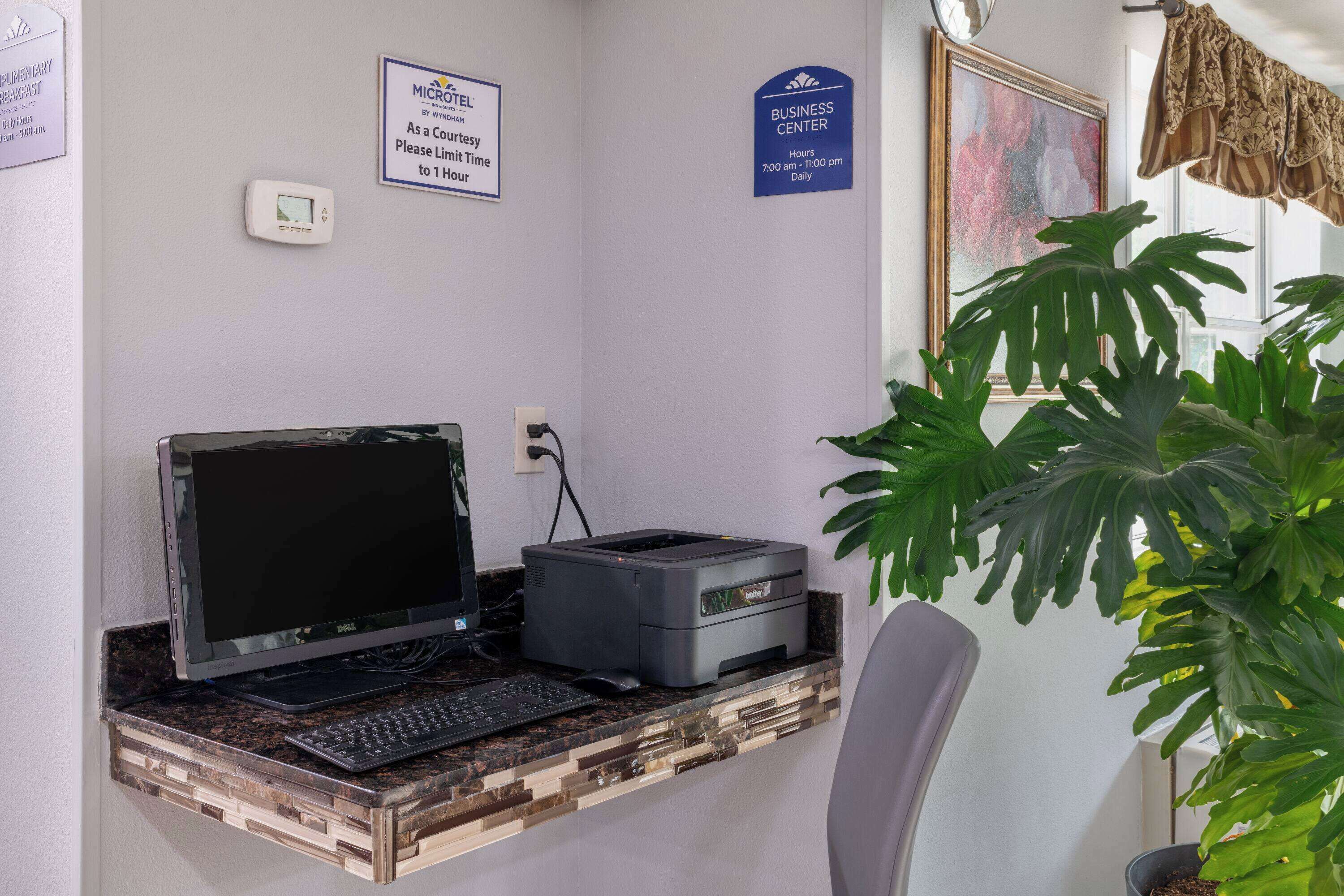 A hotel reception desk with a laptop, printer, and a computer monitor. There are signs indicating the business center hours and a courtesy limit time.