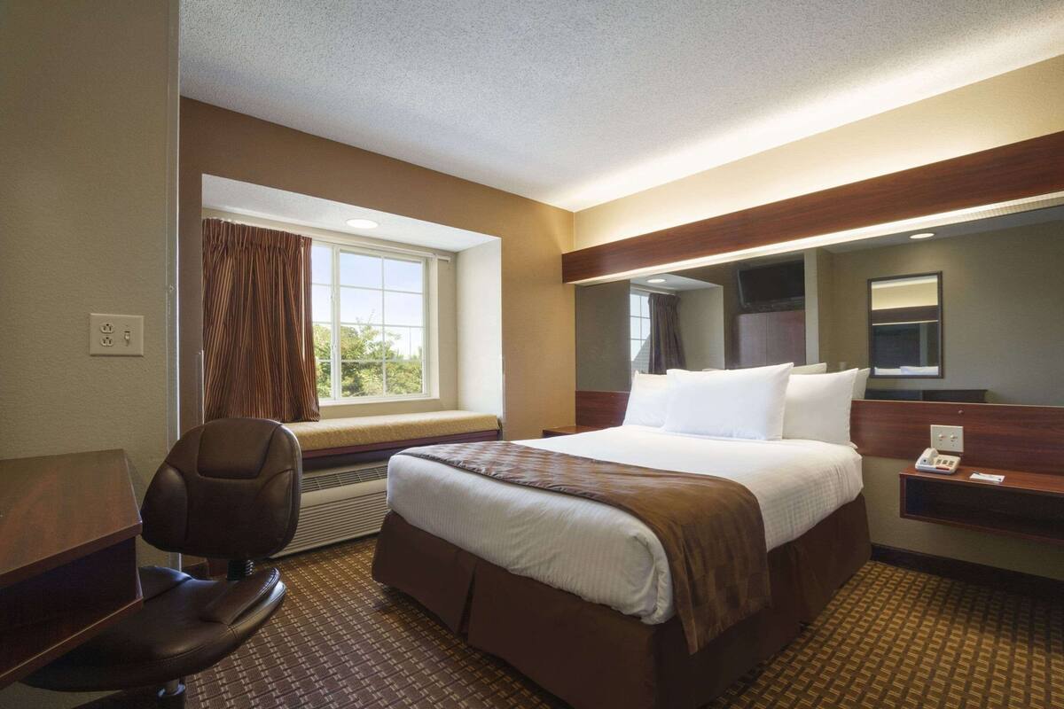 Una habitación de hotel con cama, escritorio, silla, ventana con cortinas, espejo y teléfono.