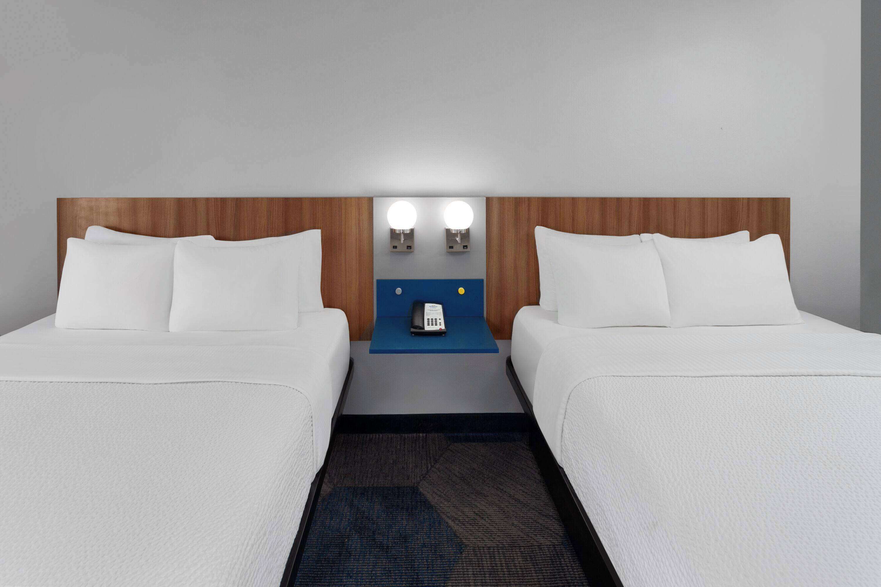 A hotel room with two beds, a wooden headboard, a blue nightstand with a phone, and two wall-mounted lights.