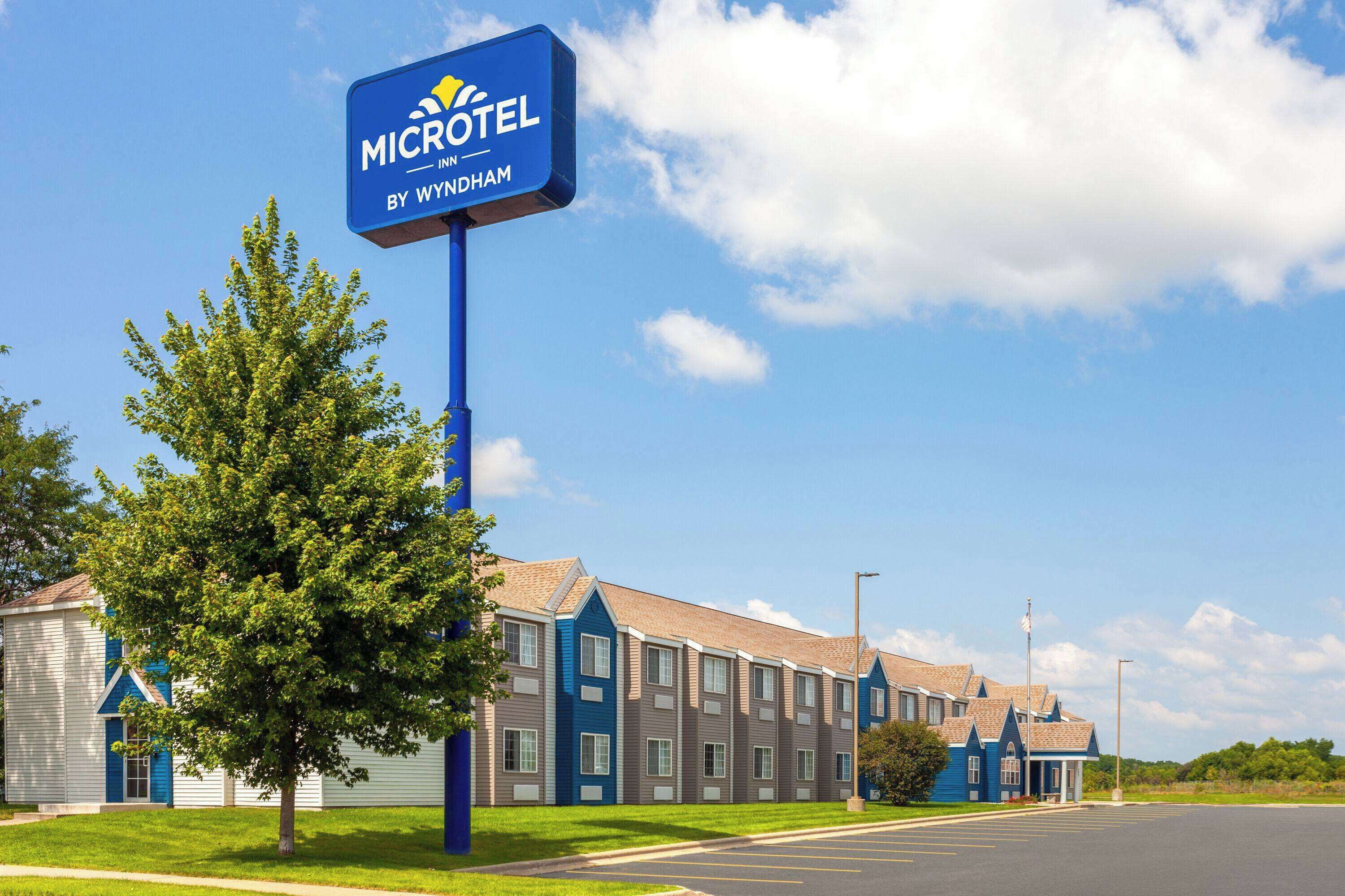 Un hotel Microtel Inn by Wyndham con un cielo azul despejado, un estacionamiento y un árbol en primer plano.
