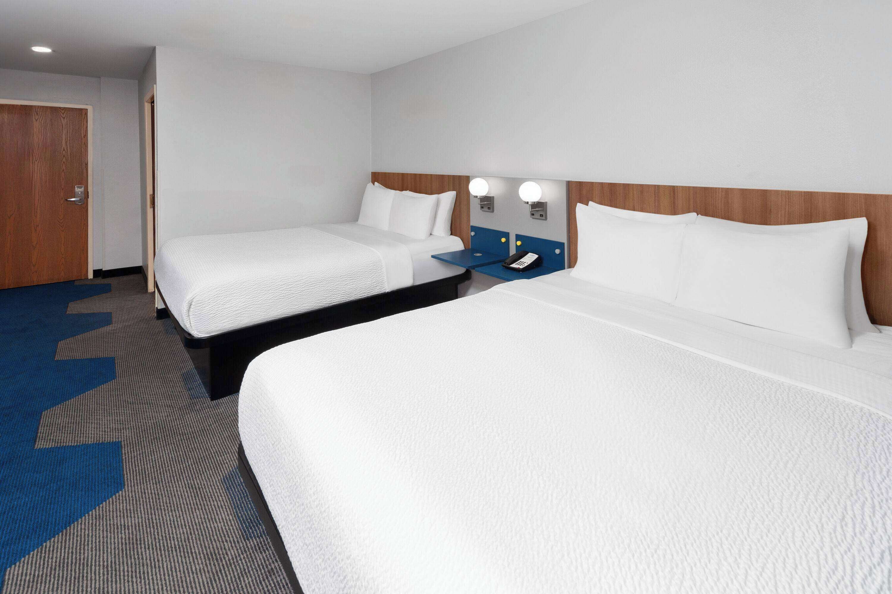 A hotel room with two single beds, a wooden headboard, bedside tables with lamps, and a blue carpet with a large 'M' pattern.