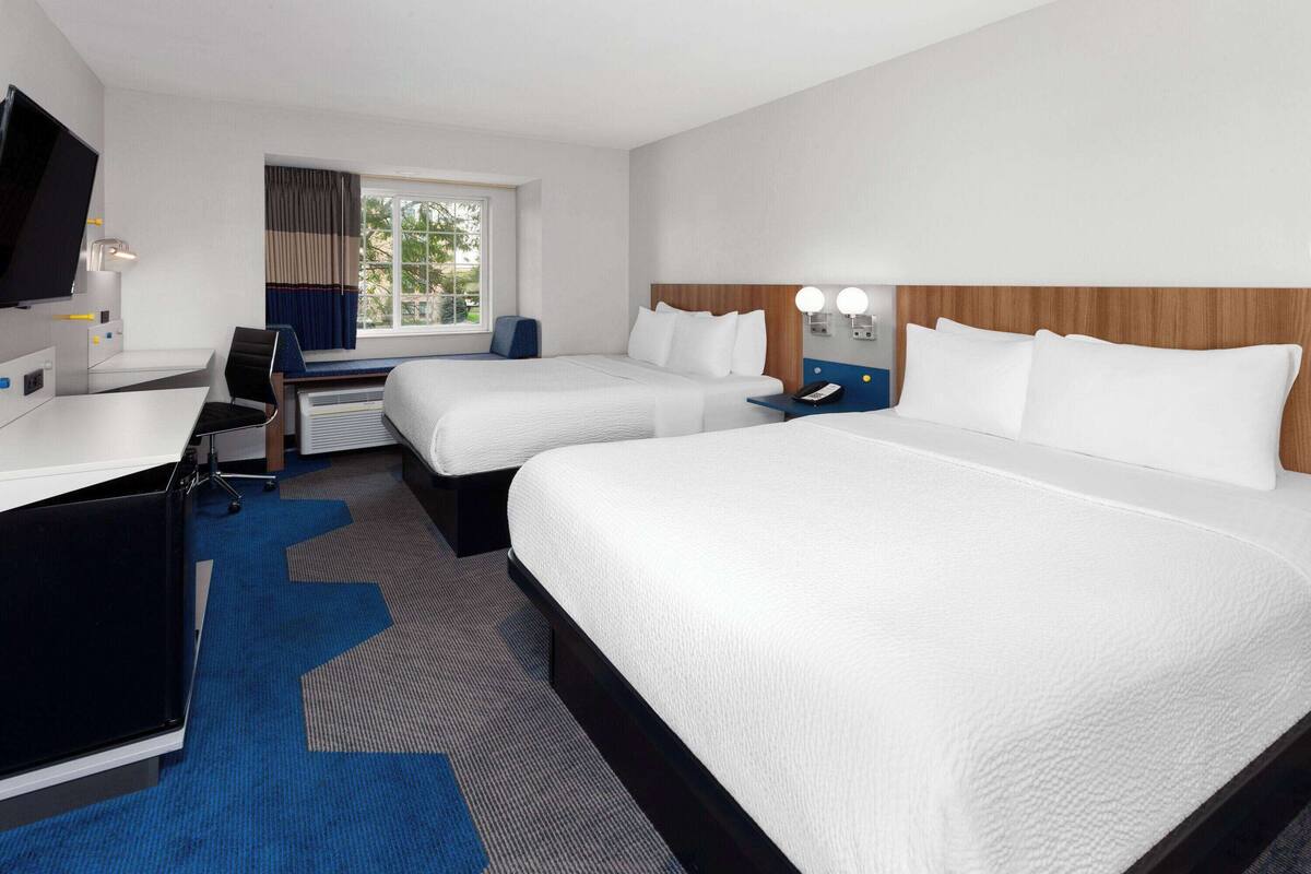 Habitación de hotel con dos camas, un escritorio, una silla, un televisor y un ventanal con cortinas.