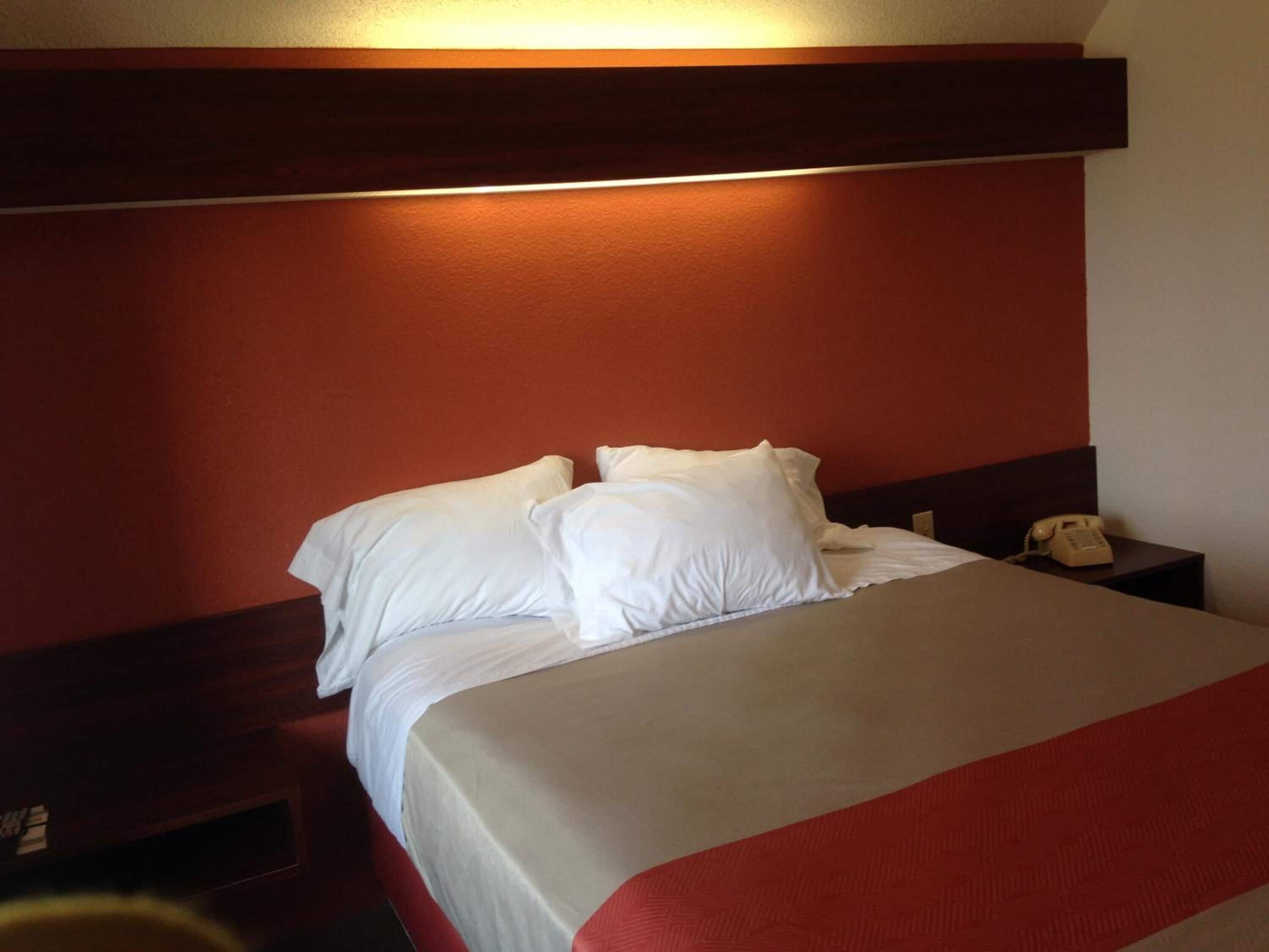 Habitación de hotel con una cama, almohadas blancas, cabecera de madera, mesita de noche con un teléfono y una alfombra roja.