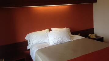 Habitación de hotel con una cama, almohadas blancas, cabecera de madera, mesita de noche con un teléfono y una alfombra roja.