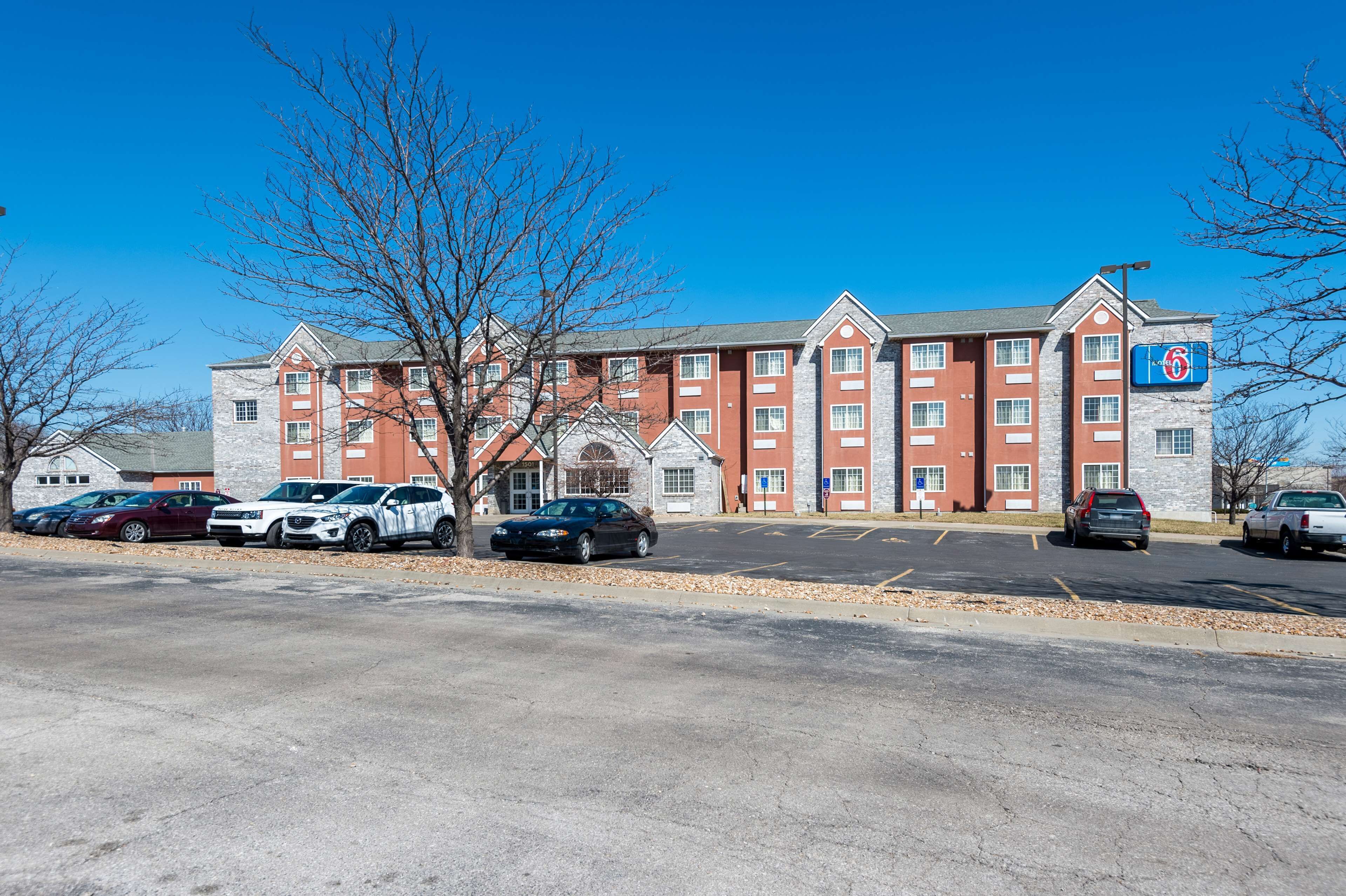 A multi-story hotel with a brick facade, a parking lot with several cars, and a clear blue sky.