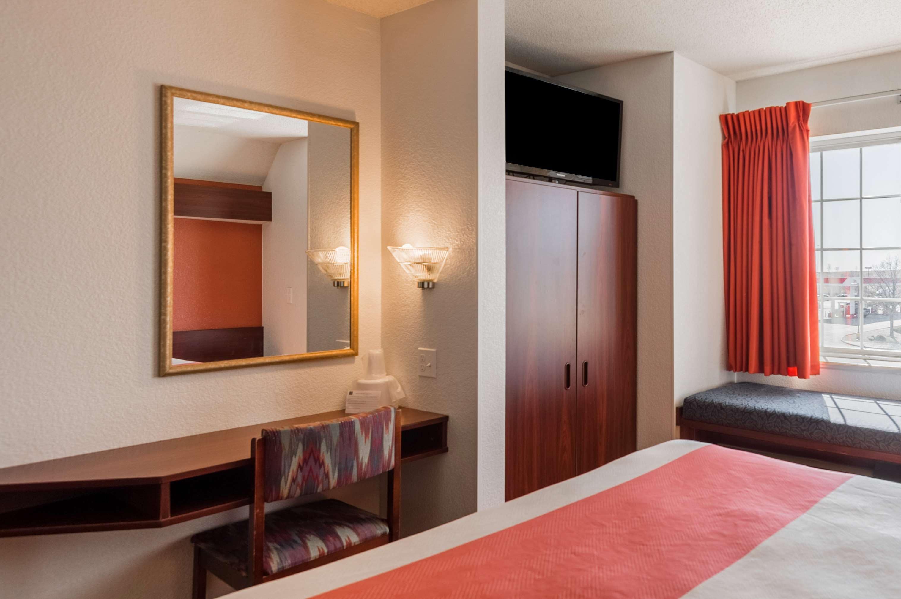 Ein Hotelzimmer mit Bett, Schreibtisch mit Stuhl, Kleiderschrank, Fernseher und einem Fenster mit Vorhängen.