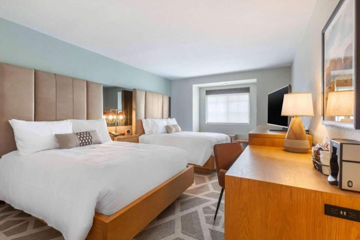 A hotel room with two beds, a desk, a TV, and a window.