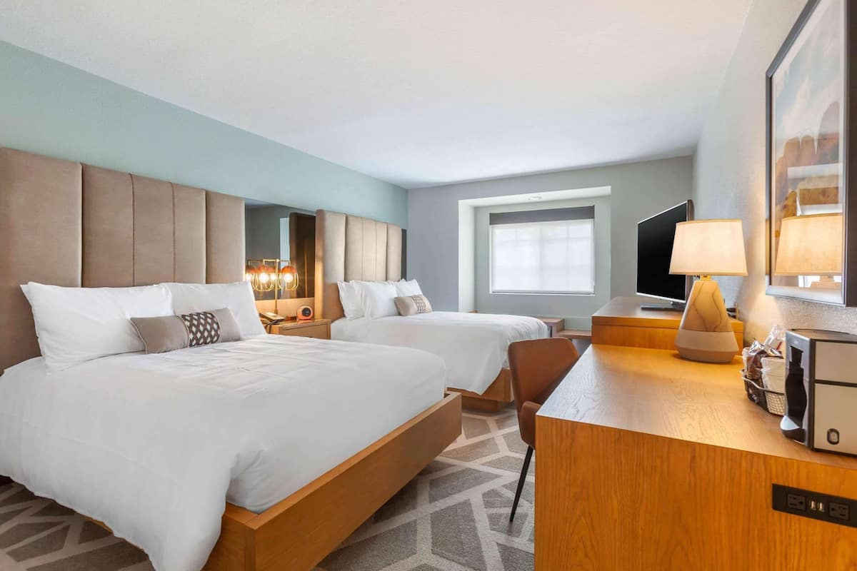 A hotel room with two beds, a desk, a TV, and a window.