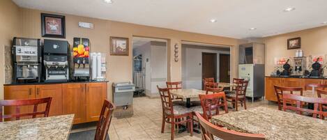 A hotel dining area with granite tables and wooden chairs, equipped with a coffee machine, juice dispenser, and a refrigerator.