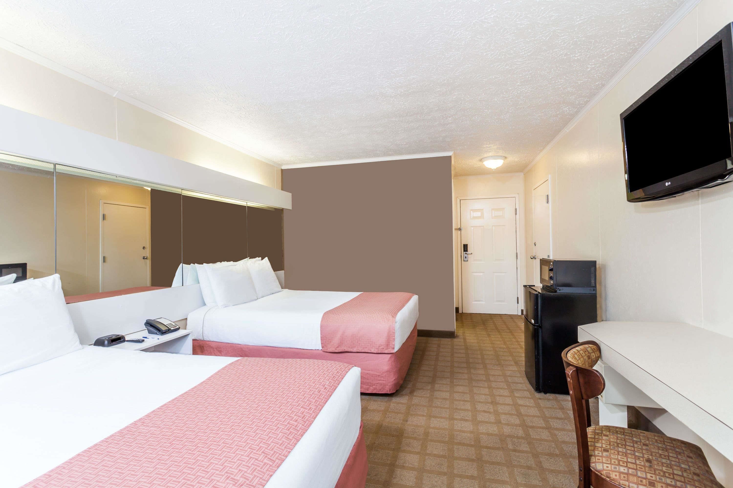 A hotel room with two beds, a desk, a television, and a small kitchenette.