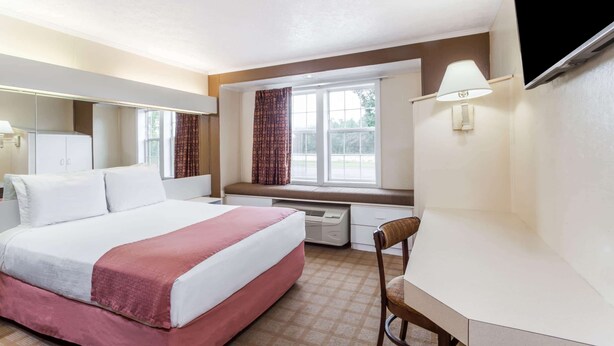 A hotel room with a bed, a desk, a chair, a window with curtains, a flat-screen TV mounted on the wall, and a wall-mounted lamp.