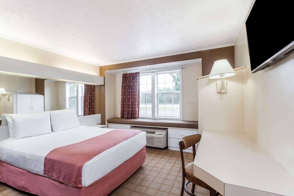 A hotel room with a bed, a desk, a chair, a window with curtains, a flat-screen TV mounted on the wall, and a wall-mounted lamp.