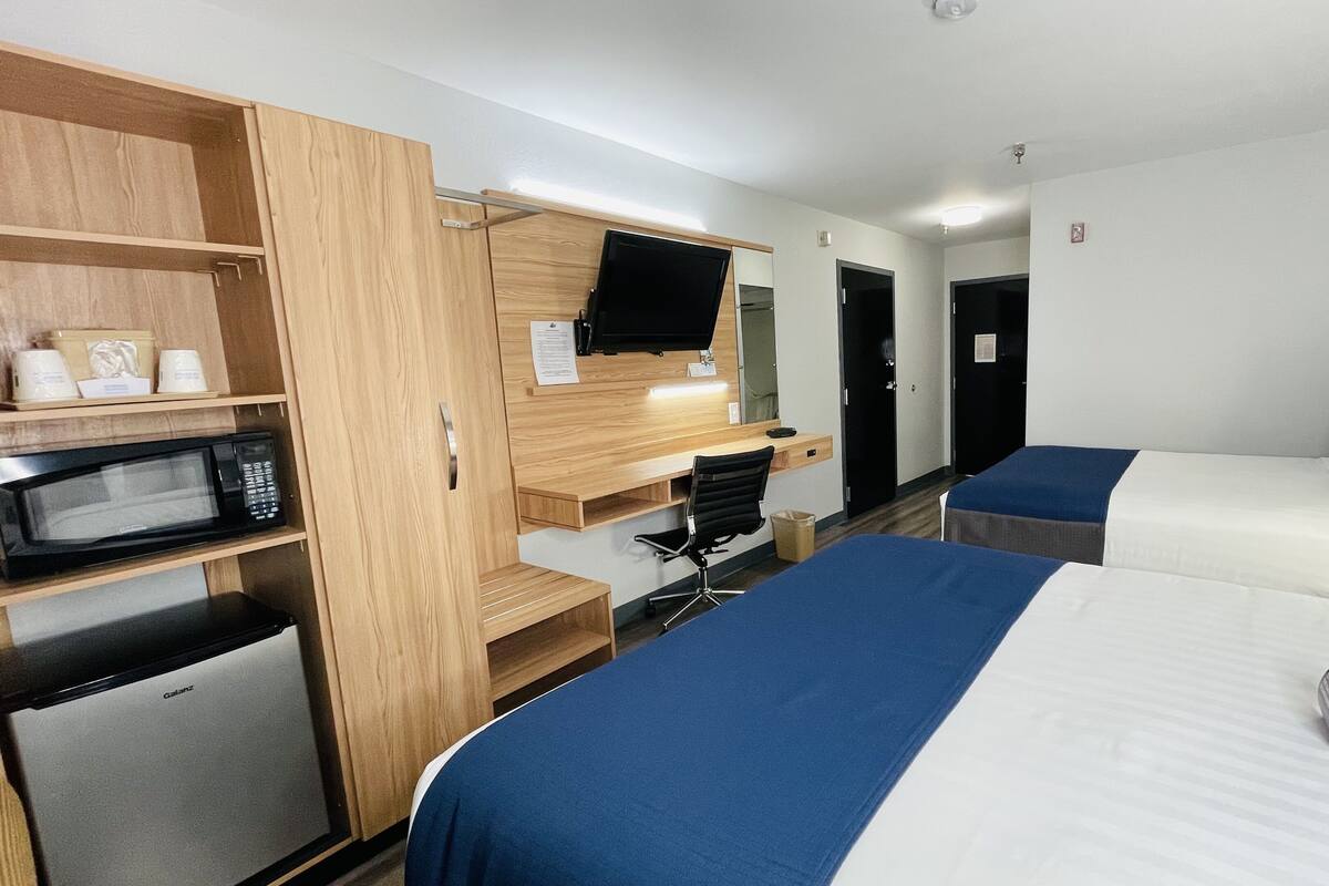 A hotel room with a kitchenette, a bed with blue and white bedding, a desk with a computer, and a TV mounted above the desk.