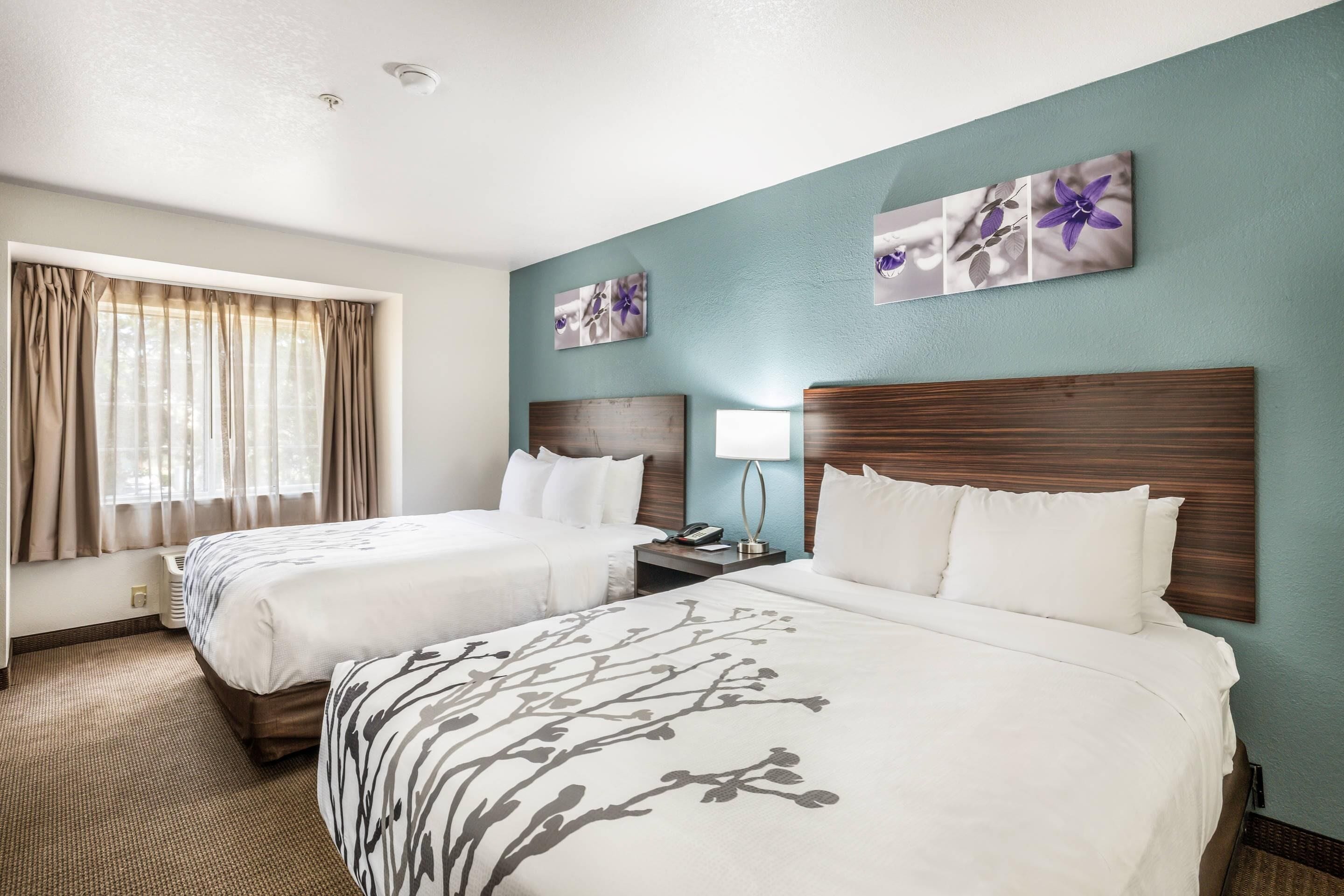 A hotel room with two beds, a window with curtains, a wall-mounted artwork, and a bedside table with a lamp.