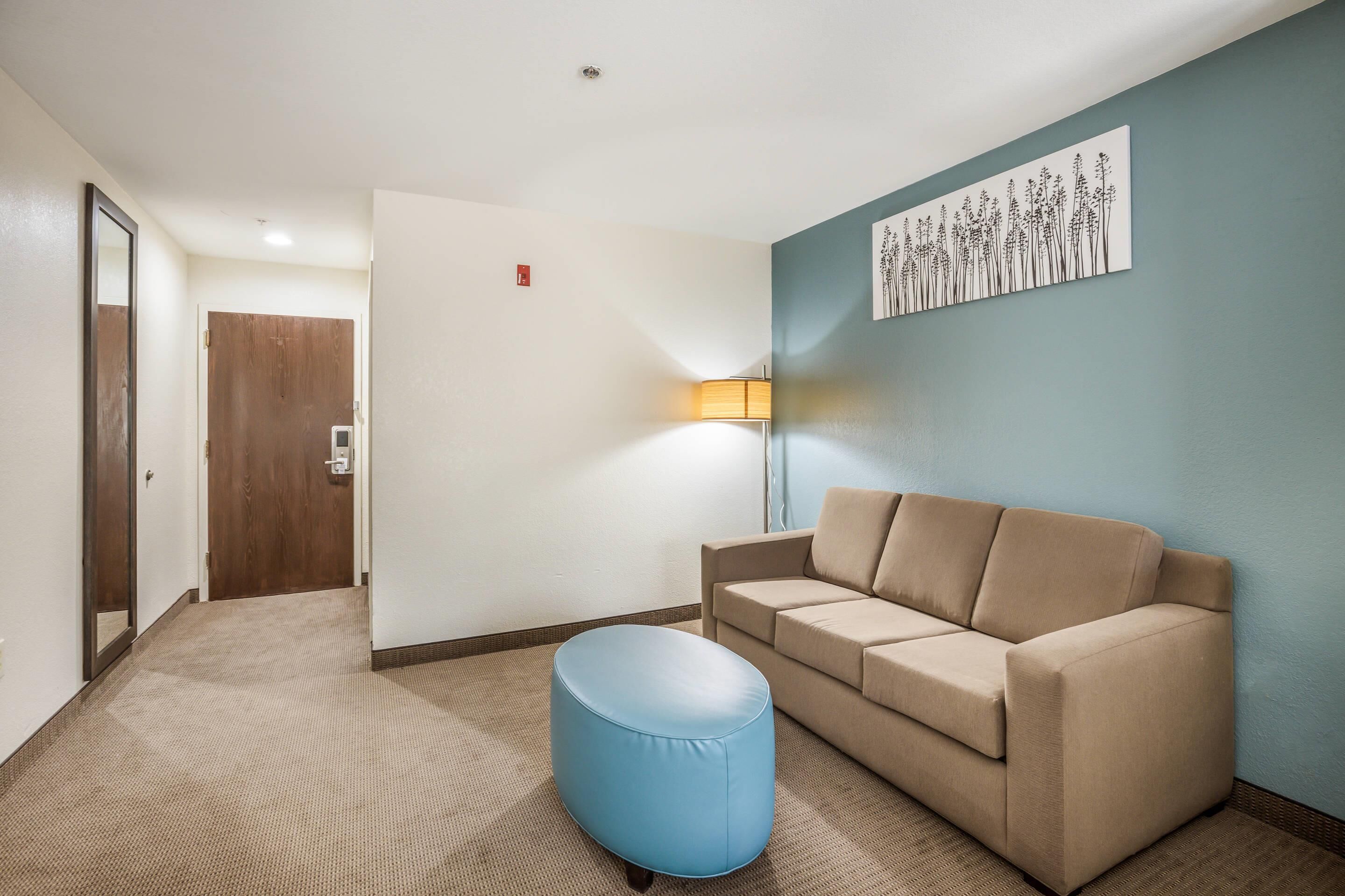 A hotel room with a beige sofa, a blue ottoman, a wall-mounted lamp, and a door with a mirror.