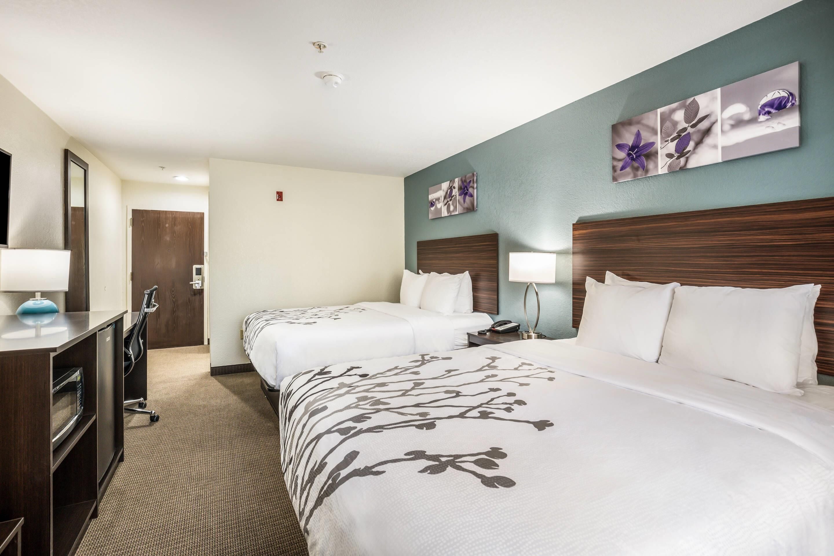 A hotel room with two beds, a desk, a mirror, and a wall-mounted artwork.