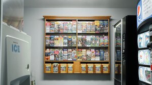 A vending machine with various snacks and drinks, a refrigerator with a 'Ice' sign, and a wall with a display of magazines and books.