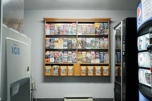 A vending machine with various snacks and drinks, a refrigerator with a 'Ice' sign, and a wall with a display of magazines and books.