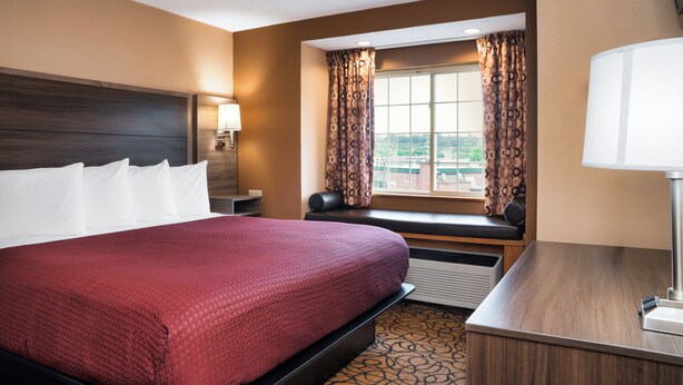 A hotel room with a large bed, a desk with a lamp, a window with curtains, and a view of the outside.